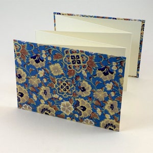 Puede incluir: Un pequeño libro de color azul y blanco con estampado floral y un interior de color crema. El libro está cerrado y de pie sobre una superficie blanca.