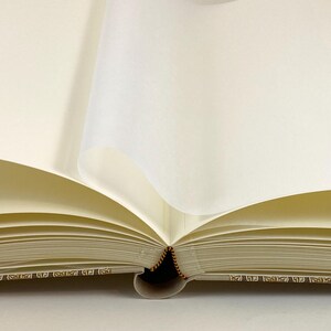 Peut inclure: Un livre ouvert avec des pages de couleur cr&egrave;me. Les pages sont l&eacute;g&egrave;rement incurv&eacute;es, avec quelques pages retourn&eacute;es. La reliure est visible et les bords des pages pr&eacute;sentent un motif d&eacute;coratif. Le livre est sur une surface blanche.