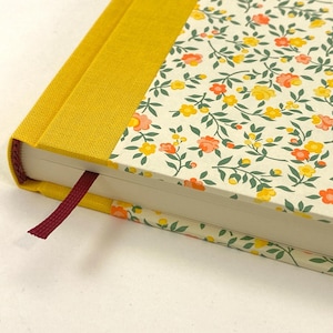Könnte beinhalten: Ein Hardcover-Journal mit floralem Muster und einem senfgelben Rücken. Das Cover hat einen cremefarbenen Hintergrund mit orangefarbenen und gelben Blumen und grünen Blättern. Ein burgunderfarbenes Lesezeichenband ist sichtbar.