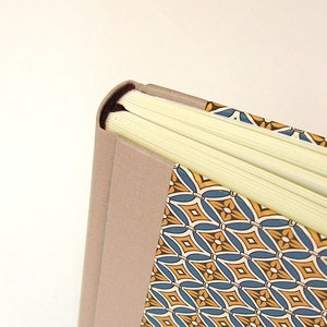 Puede incluir: Primer plano del lomo de un libro de tapa dura con una cubierta de lino beige y un dise&ntilde;o estampado de colores. El libro est&aacute; cerrado y las p&aacute;ginas son visibles.