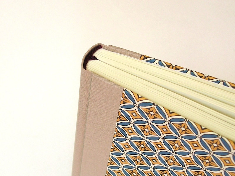 Puede incluir: Primer plano del lomo de un libro de tapa dura con una cubierta de lino beige y un dise&ntilde;o estampado de colores. El libro est&aacute; cerrado y las p&aacute;ginas son visibles.
