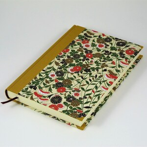 May include: A cream-coloured journal with a floral pattern on the cover. The cover features red, black, and green flowers and leaves on a cream background. The spine of the journal is a solid gold colour.
