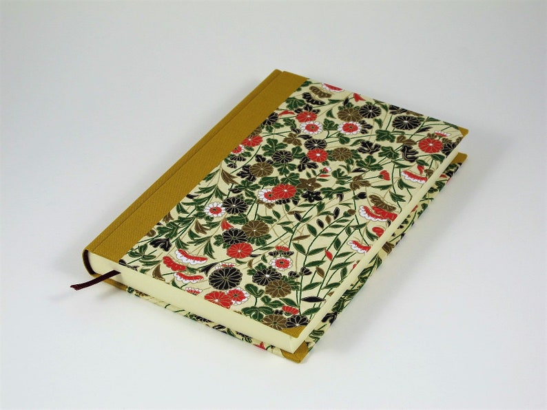 May include: A cream-coloured journal with a floral pattern on the cover. The cover features red, black, and green flowers and leaves on a cream background. The spine of the journal is a solid gold colour.