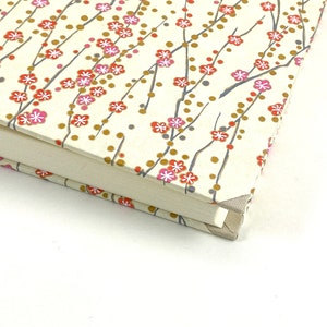 May include: Close-up of the corner of a cream-colored journal with a floral pattern. The pattern features small pink and red flowers with gold accents on thin gray branches.