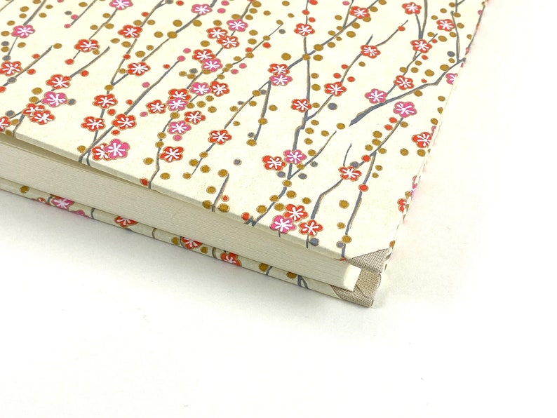 May include: Close-up of the corner of a cream-colored journal with a floral pattern. The pattern features small pink and red flowers with gold accents on thin gray branches.