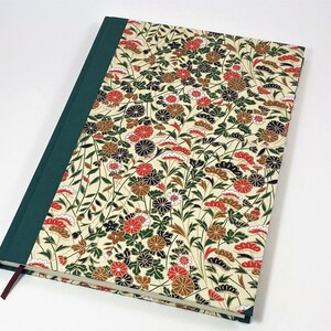 Op de afbeelding: Een groen en cr&egrave;mekleurig notitieboek met een bloemenpatroon op de omslag. De bloemen zijn rood, wit en zwart. Het notitieboek heeft een groene rug en een rood lint als bladwijzer.