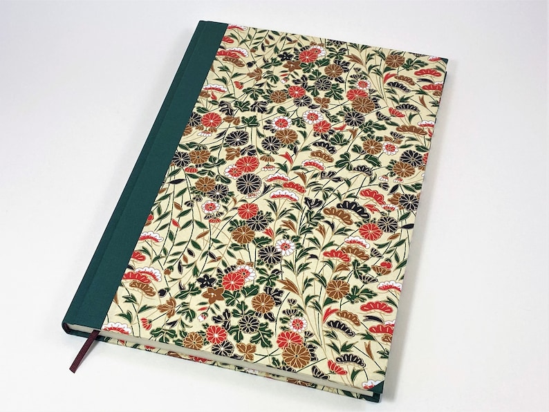 Op de afbeelding: Een groen en cr&egrave;mekleurig notitieboek met een bloemenpatroon op de omslag. De bloemen zijn rood, wit en zwart. Het notitieboek heeft een groene rug en een rood lint als bladwijzer.