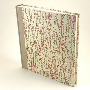 May include: A cream-colored hardcover journal with a floral pattern on the cover. The pattern features small pink flowers and brown branches on a cream background. The spine of the journal is a light brown color.