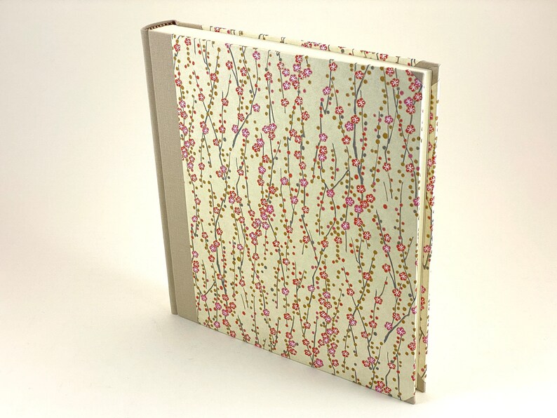 May include: A cream-colored hardcover journal with a floral pattern on the cover. The pattern features small pink flowers and brown branches on a cream background. The spine of the journal is a light brown color.