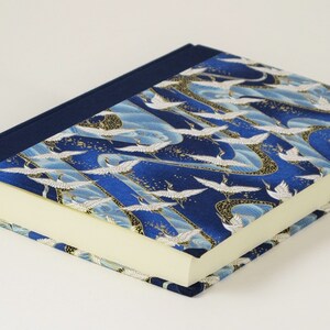 May include: A blue notebook with a white crane pattern on the cover. The notebook has a dark blue spine and a cream-colored interior.
