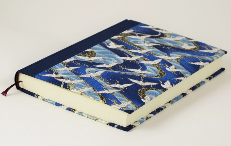 May include: A blue notebook with a white crane pattern on the cover. The notebook has a dark blue spine and a cream-colored interior.