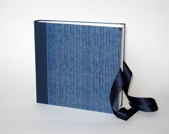 May include: A blue fabric-covered photo album with a white and blue patterned design. The album has a dark blue ribbon tied around it.