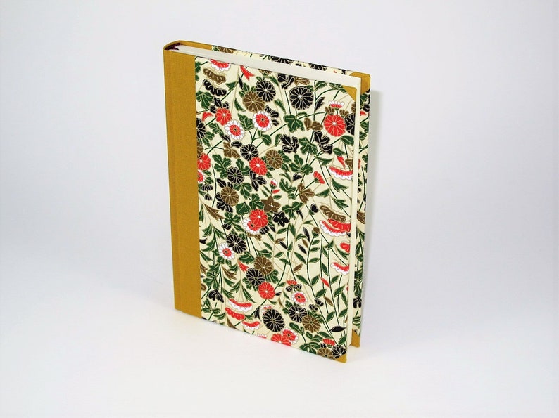 May include: A hardcover journal with a floral design on the cover. The cover features a white background with red, black, and green flowers and leaves. The spine is a solid gold colour.