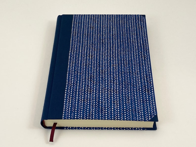 Pode incluir: Um di&aacute;rio de capa dura azul com um design estampado branco e um marcador de fita cor vinho.