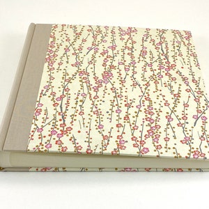 May include: A cream-colored hardcover photo album with a floral design on the front cover. The design features pink and red flowers on thin branches with small gold dots.