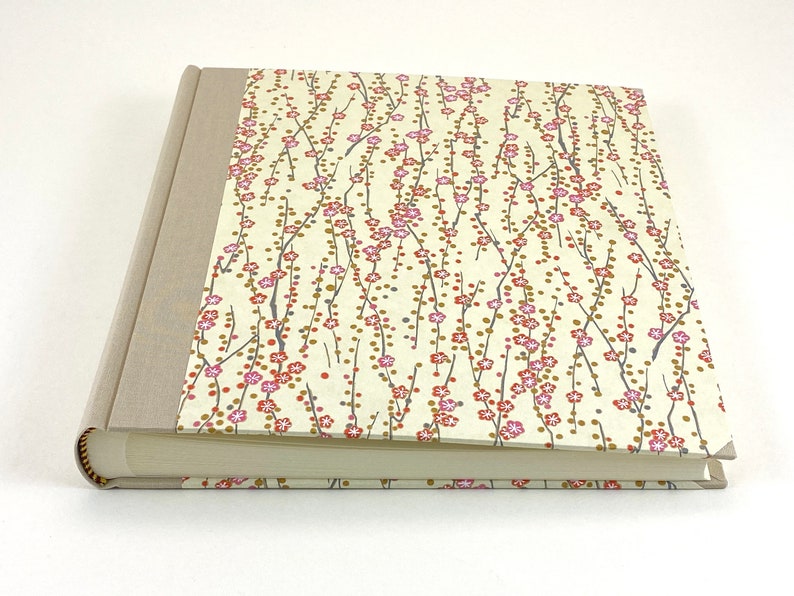 May include: A cream-colored hardcover photo album with a floral design on the front cover. The design features pink and red flowers on thin branches with small gold dots.