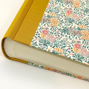 May include: A hardcover book with a mustard yellow spine and a floral pattern cover. The cover features a cream background with a repeating pattern of pink, orange, and gray flowers with green leaves. The book's pages are white.