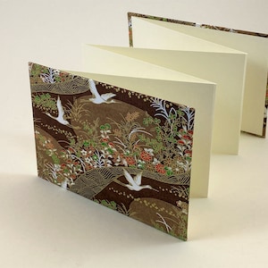May include: A brown and gold paperbound journal with a white crane pattern. The journal is folded open to reveal blank pages.