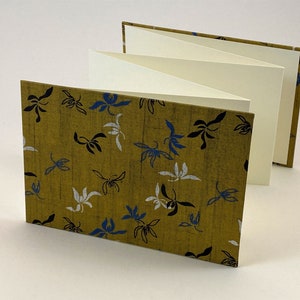 May include: A small, folded book with a mustard yellow cover featuring a repeating pattern of white and blue stylized flowers. The book is open to reveal blank white pages.