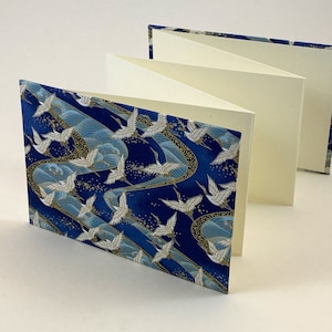 May include: A blue and gold patterned fabric-covered journal with white cranes flying over waves. The journal has multiple pages and is folded closed.