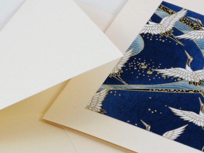 May include: A cream-colored card with a blue and gold patterned insert featuring white cranes flying against a dark blue background. The pattern includes gold dots and lines.
