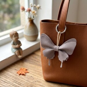 May include: A tan pebbled leather tote bag featuring a light grey butterfly-shaped charm attached to the handle with a silver metal ring. The butterfly charm is constructed from layered material with a smooth finish and a central loop attachment.