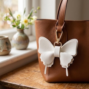 May include: A brown leather tote bag featuring a textured surface and a white butterfly-shaped charm attached to the handle with a gold-toned metal lobster clasp. The butterfly charm is constructed from layered white material with a smooth finish.