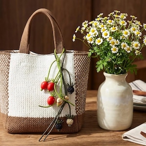 May include: A woven tote bag with a white and brown pattern, brown handles, and a decorative charm featuring red berries, gold beads, and black berries. A white vase holds a bouquet of white and yellow flowers.
