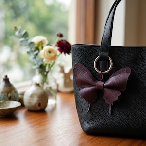 May include: A black pebbled leather tote bag featuring a dark purple butterfly-shaped charm attached to a silver-toned metal ring. The bag has a structured vertical design with a single visible handle and a textured surface finish.