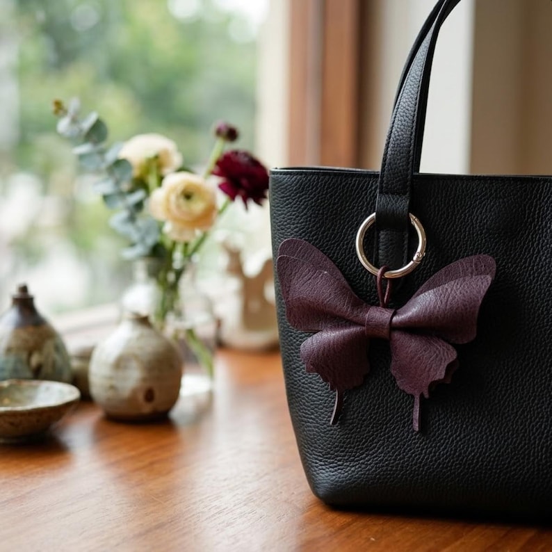 May include: A black pebbled leather tote bag featuring a dark purple butterfly-shaped charm attached to a silver-toned metal ring. The bag has a structured vertical design with a single visible handle and a textured surface finish.
