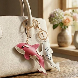 May include: A cream-coloured handbag features a gold-toned metal keychain with two charms. One charm is a pink and white whale-shaped pendant, and the other is a white fin-shaped tag with the black text "Hairly" and a small heart symbol printed on it.