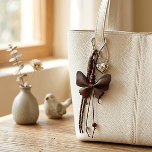 May include: A cream-coloured textured leather tote bag featuring a brown leather butterfly charm attached to a silver-toned metal clasp. The charm includes a small silver heart pendant, brown leather cords, and small wooden and metallic beads.