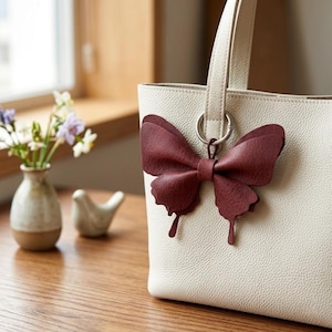 May include: A cream-coloured tote bag with a textured surface featuring a large, dark red butterfly-shaped charm attached to a silver-toned metal ring. The butterfly charm is made of a layered material with a matte finish and distinct wing shapes.