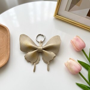 May include: A metallic gold butterfly-shaped keyring featuring a textured surface and a silver-toned metal ring attachment. The butterfly design has layered wings with a central band, creating a dimensional appearance for this accessory.
