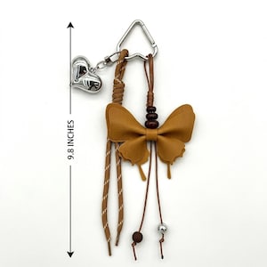 May include: A brown butterfly-shaped charm attached to a silver-toned heart charm and a triangular metal ring. The accessory features brown cords, dark brown beads, and a silver bead, with a total length of 24.89 cm as indicated by the arrow.