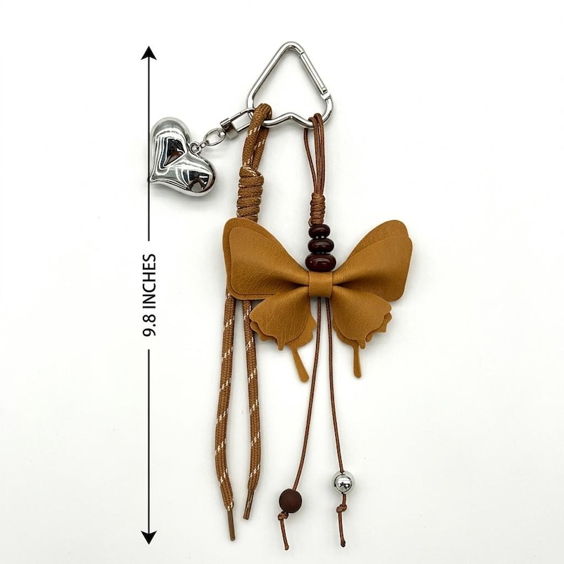 May include: A brown butterfly-shaped charm attached to a silver-toned heart charm and a triangular metal ring. The accessory features brown cords, dark brown beads, and a silver bead, with a total length of 24.89 cm as indicated by the arrow.