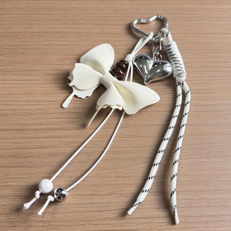 May include: A keyring featuring a white faux leather bow, a silver-toned heart charm, and a heart-shaped ring. The design includes white cords with black flecks, dark brown beads, and two small spherical beads in white and metallic silver finishes.