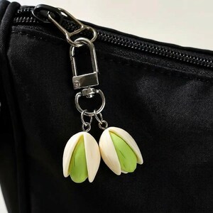 May include: A silver-coloured keyring with a lobster clasp and two pistachio-shaped charms. The charms feature a light green pistachio nut encased in a white shell. The keyring is attached to a black bag.