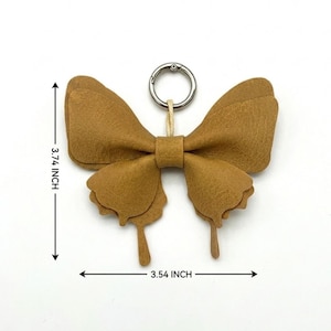 May include: A tan butterfly-shaped keyring made of layered material with a silver metal ring. The item measures 9.5 cm in height and 9.0 cm in width, as indicated by the black arrows and text labels on the image.