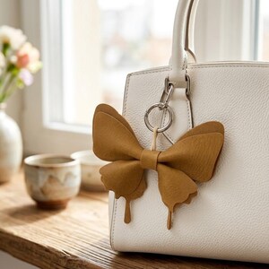 May include: A white textured handbag featuring a mustard-coloured butterfly-shaped charm attached to the silver metal hardware. The charm is made of a smooth, matte material and hangs from a circular ring connected to the bag's handle base.