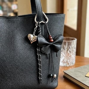May include: A black textured tote bag featuring a silver-toned heart charm and a black butterfly-shaped accessory. The butterfly is accented with three small brown wooden beads and two dangling black cords, each finished with a single bead at the end.