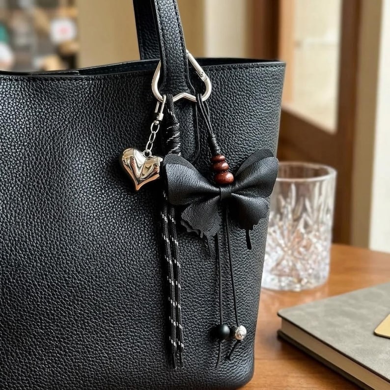 May include: A black textured tote bag featuring a silver-toned heart charm and a black butterfly-shaped accessory. The butterfly is accented with three small brown wooden beads and two dangling black cords, each finished with a single bead at the end.
