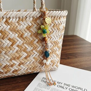 Puede incluir: Un bolso de paja tejido con asas trenzadas presenta un adorno decorativo. El adorno consta de un anillo dorado, una etiqueta redonda de metal dorado con texto, una cuenta de flor verde translúcida y varias cuentas de madera y de colores.