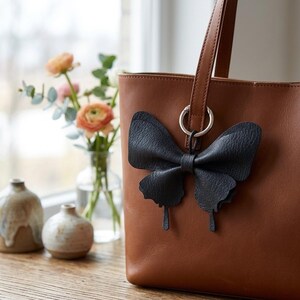 May include: A tan leather tote bag featuring a dark navy blue butterfly-shaped charm attached to the handle by a silver metal ring. The butterfly charm has a textured surface and is positioned on the front exterior of the bag.