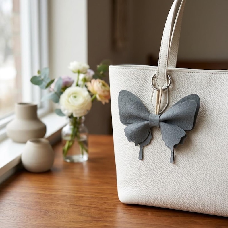May include: A white textured leather tote bag featuring a grey felt butterfly-shaped charm attached to the handle ring. The bag has a structured silhouette with a single visible handle and a smooth, clean finish on the front panel.