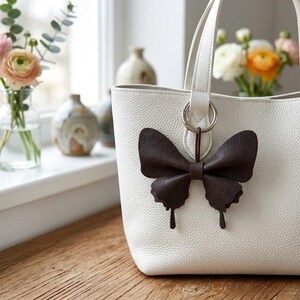 May include: A white textured leather tote bag featuring a dark brown leather butterfly-shaped charm attached to the silver-toned ring of the handle. The bag has a structured shape with visible grain and a clean, minimalist design aesthetic.
