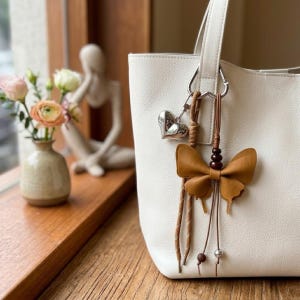 May include: A white tote bag featuring a silver-toned metal heart charm and a brown leather butterfly-shaped bag charm. The butterfly charm is accented with small dark brown beads and thin leather cords that hang down with silver-toned metal beads.