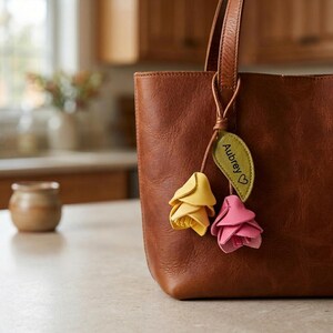 May include: A brown leather tote bag featuring a decorative charm attached to the handle. The charm consists of a green leaf-shaped tag engraved with the name Aubrey and a heart symbol, accompanied by two layered flower shapes in yellow and pink.