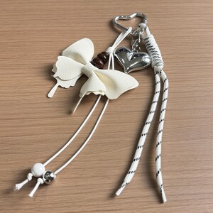 May include: A keyring featuring a white faux leather bow, a metallic silver heart charm, and a heart-shaped ring. It includes white cords with black dashed patterns, a single white bead, a metallic silver bead, and a dark brown bead accent.