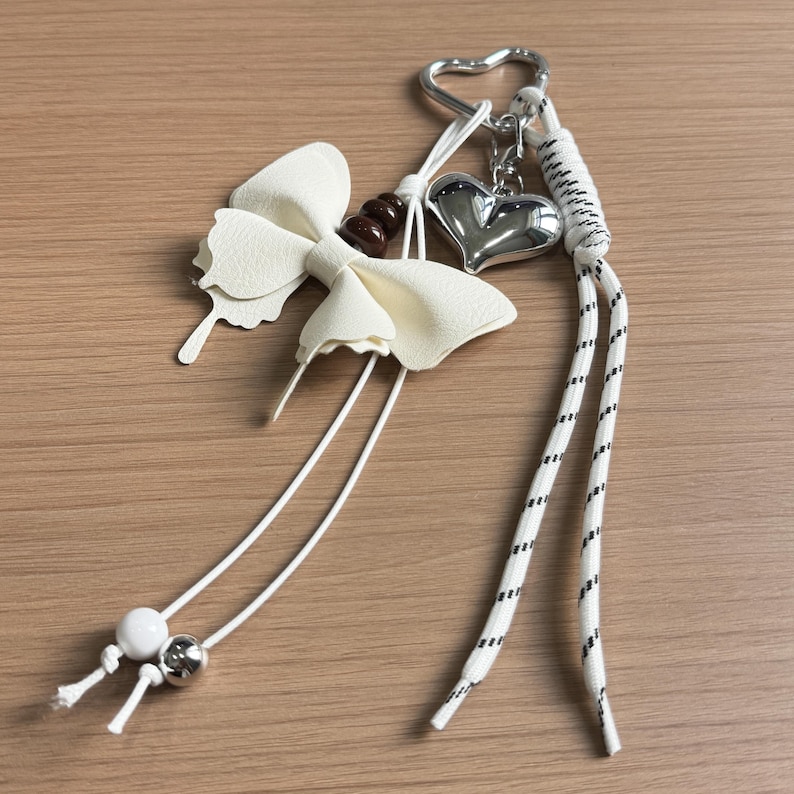May include: A keyring featuring a white faux leather bow, a metallic silver heart charm, and a heart-shaped ring. It includes white cords with black dashed patterns, a single white bead, a metallic silver bead, and a dark brown bead accent.